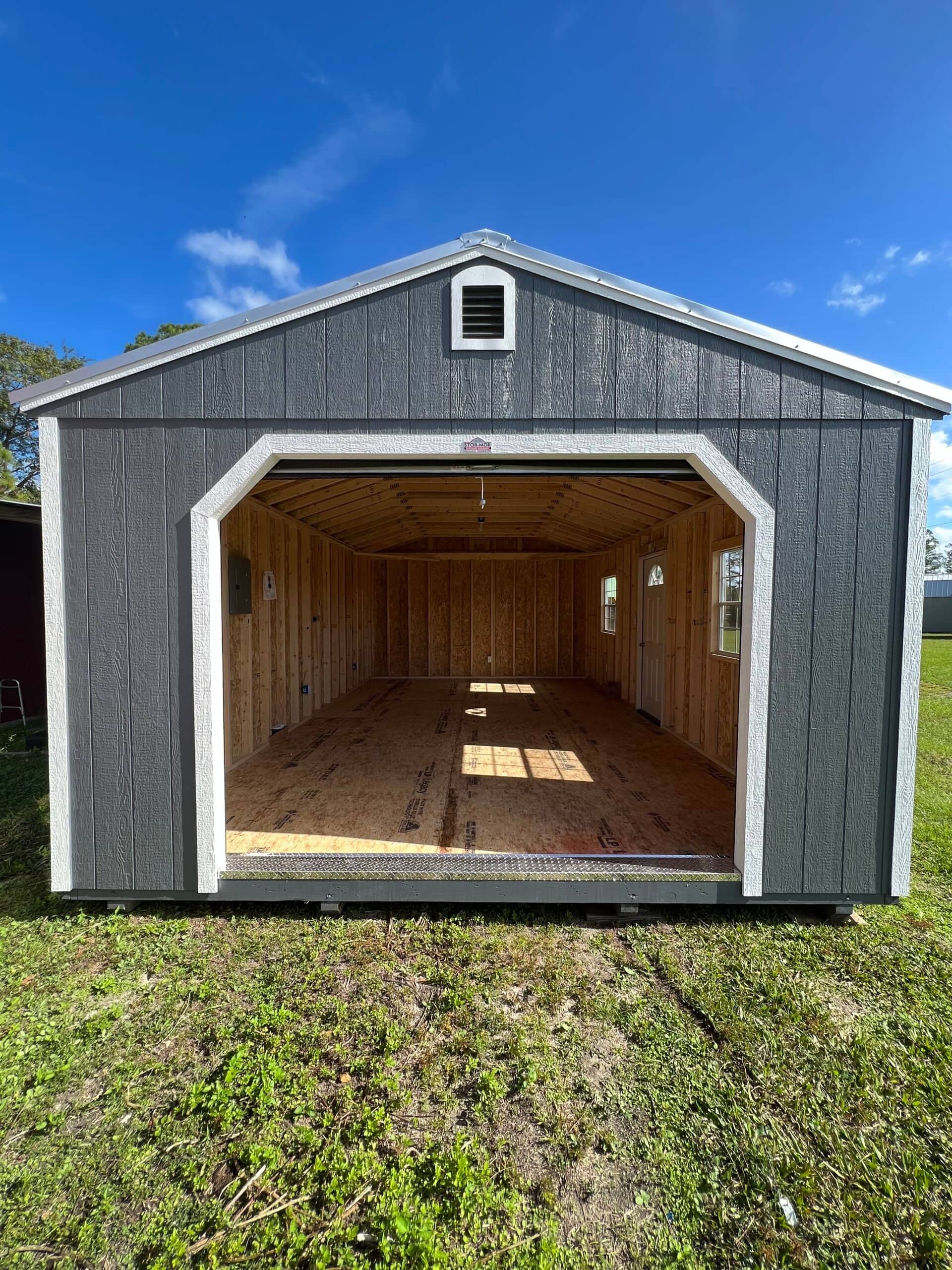 Front view looking into a dark grey high-barn garage storage shed with white trim, an open roll-up front door, unfinished wood stud interior, and LP subflooring in Chiefland, FL.