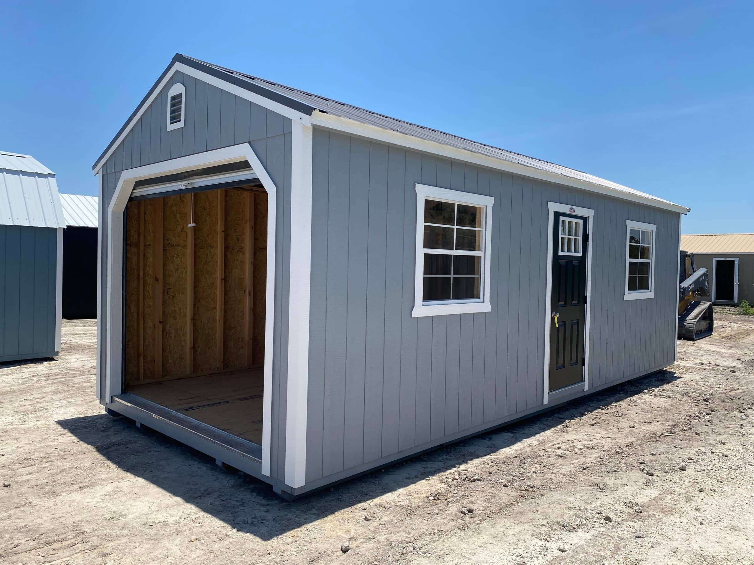 Exterior view of a grey high-barn portable garage storage shed with white trim, open roll-up front garage door, dark side door, and two grid windows in Leesburg, FL.