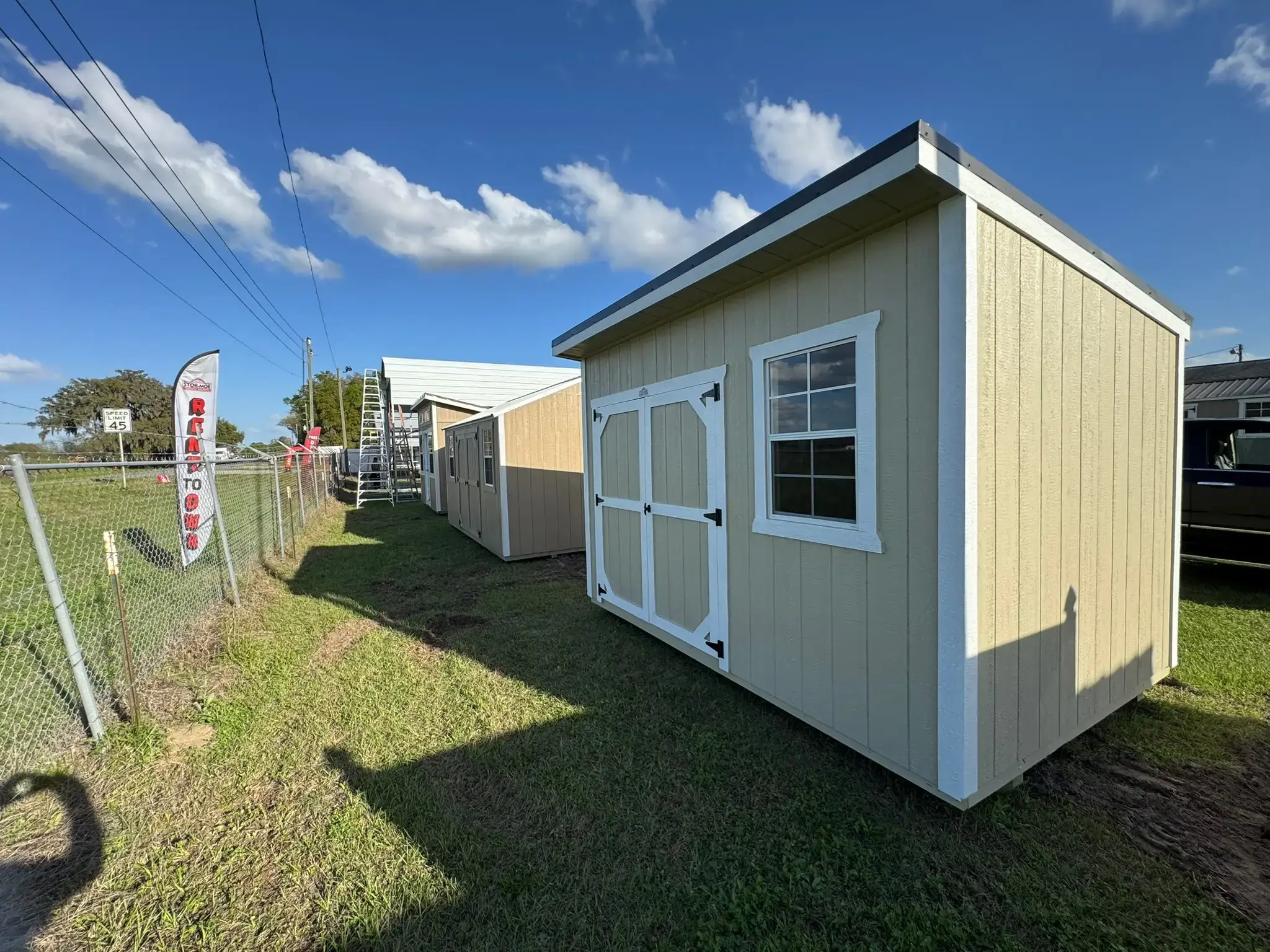 Tan portable storage building with white trim, double doors, and a single framed window sitting on grass in a outdoor sales lot in Leesburg, FL.