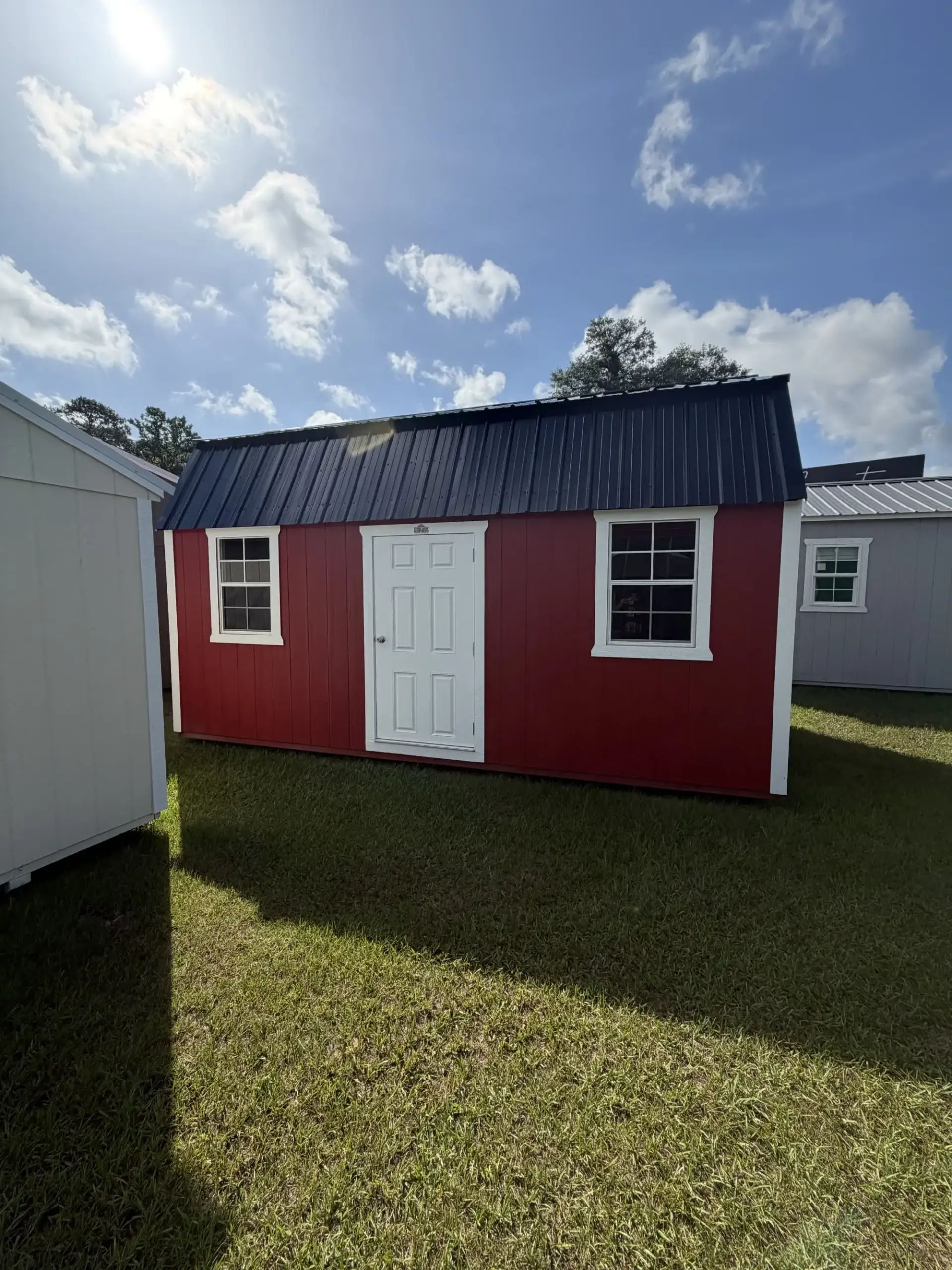 Classic red lofted barn shed with white trim, black metal gambrel roof, white paneled entry door, and two grid windows sitting on grass under a sunny sky in Ocala, FL.
