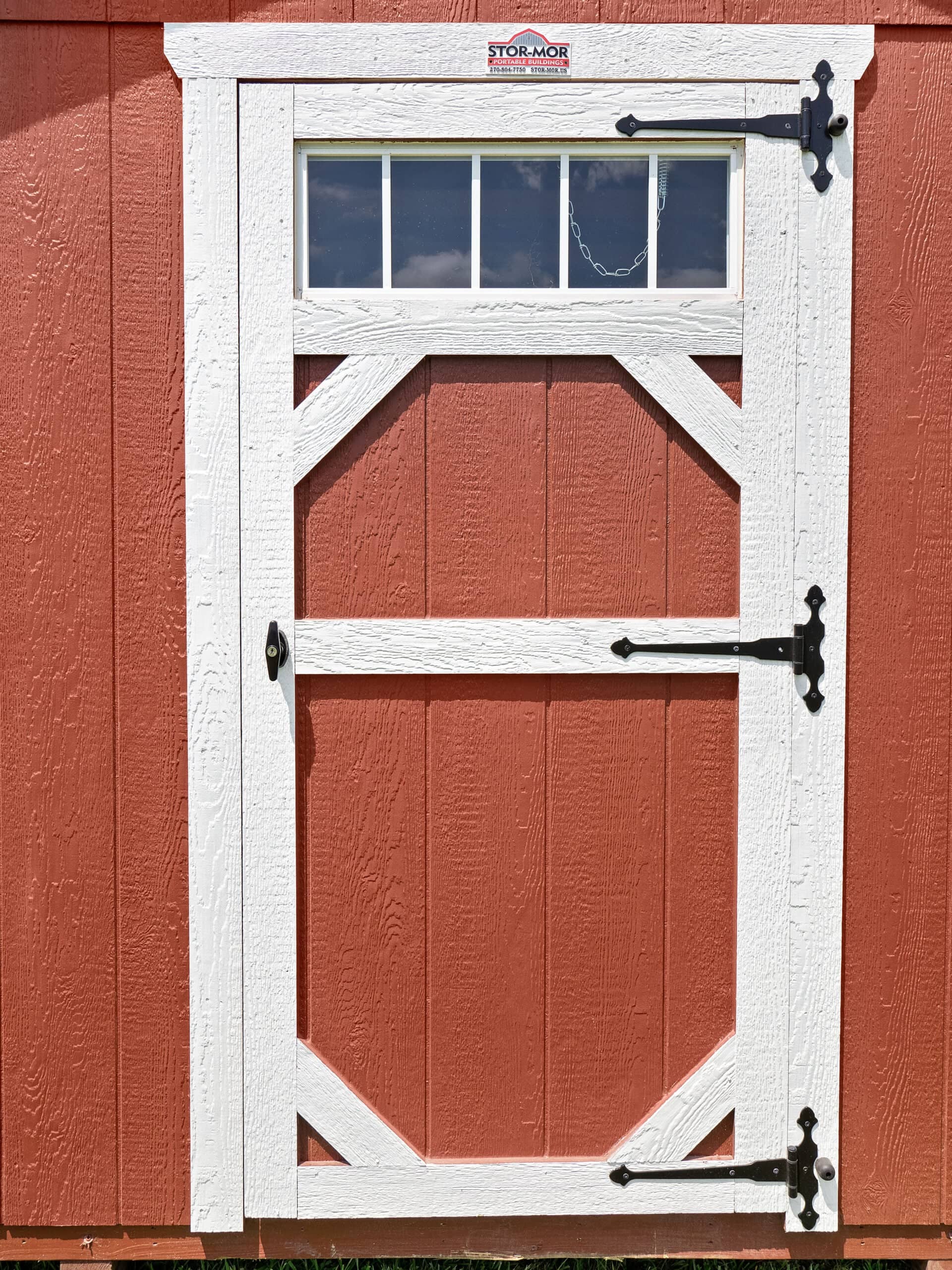 Close-up view of a custom single shed transom door featuring white trim, a multi-pane upper window, black decorative T-hinges, and a red wood-paneled background in Tavares, FL.