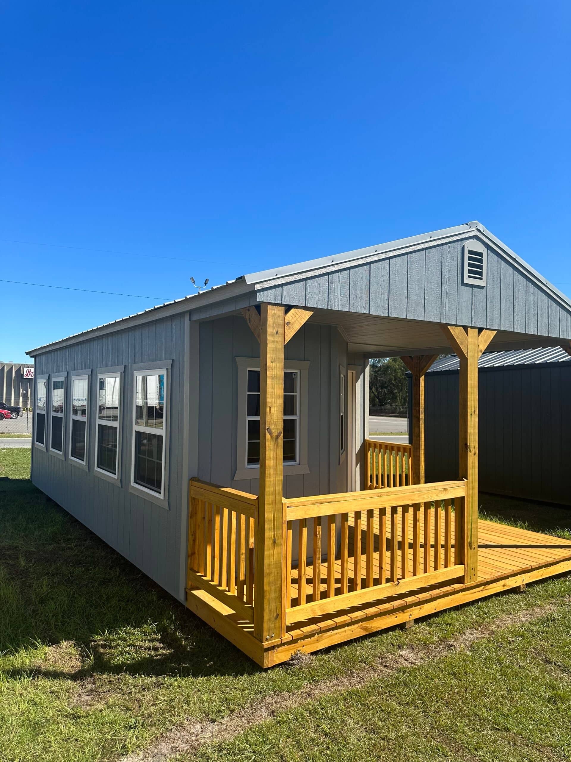 Gray wooden cabin shed with a large front wood porch, metal gable roof, white-framed grid windows, and natural wood railings in Leesburg, FL.