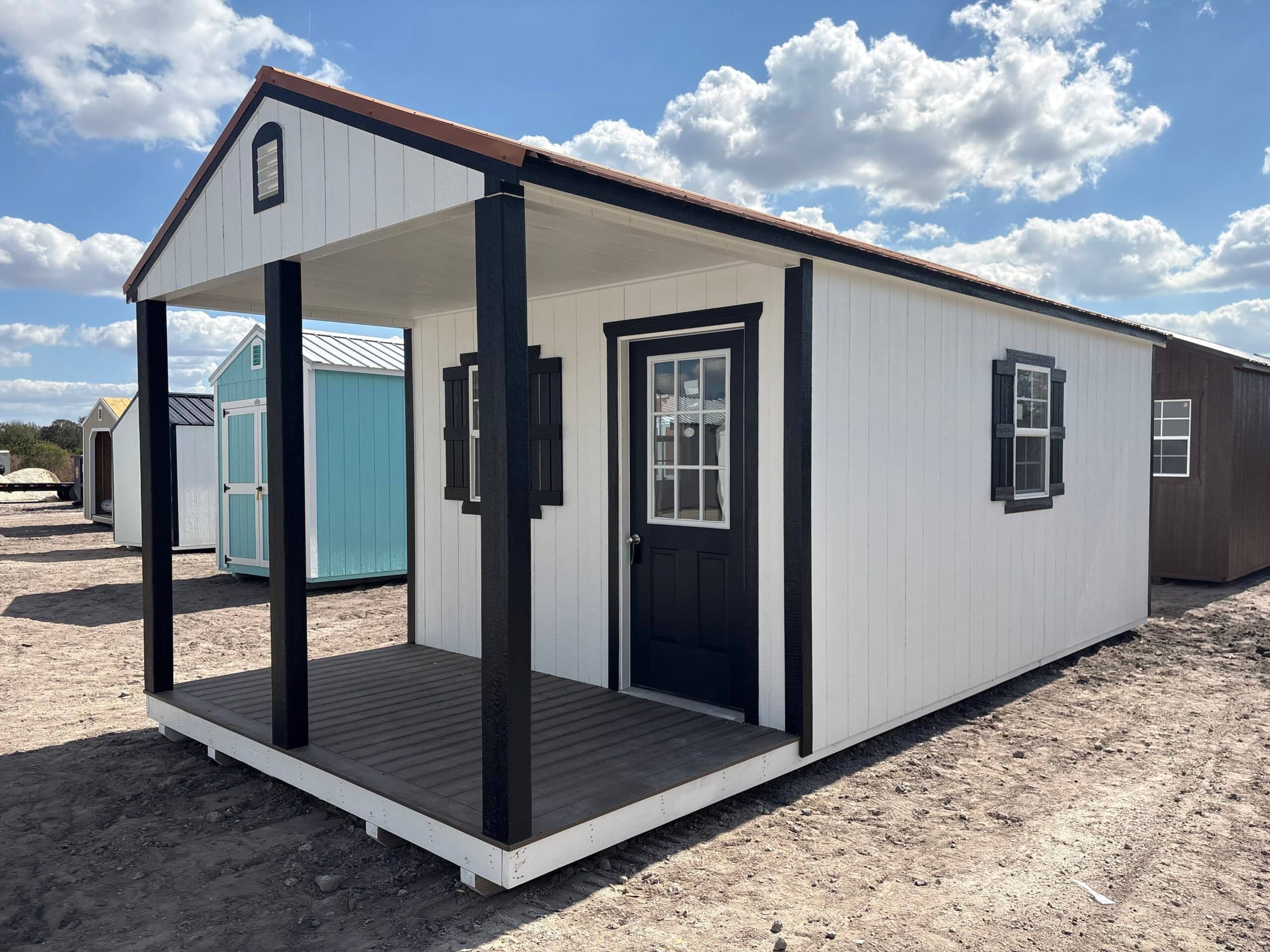 White wooden cabin utility shed featuring bold black porch columns, black trim and shutters, glass-paneled front door, front porch deck, and side window under a blue sky in Inverness, FL.