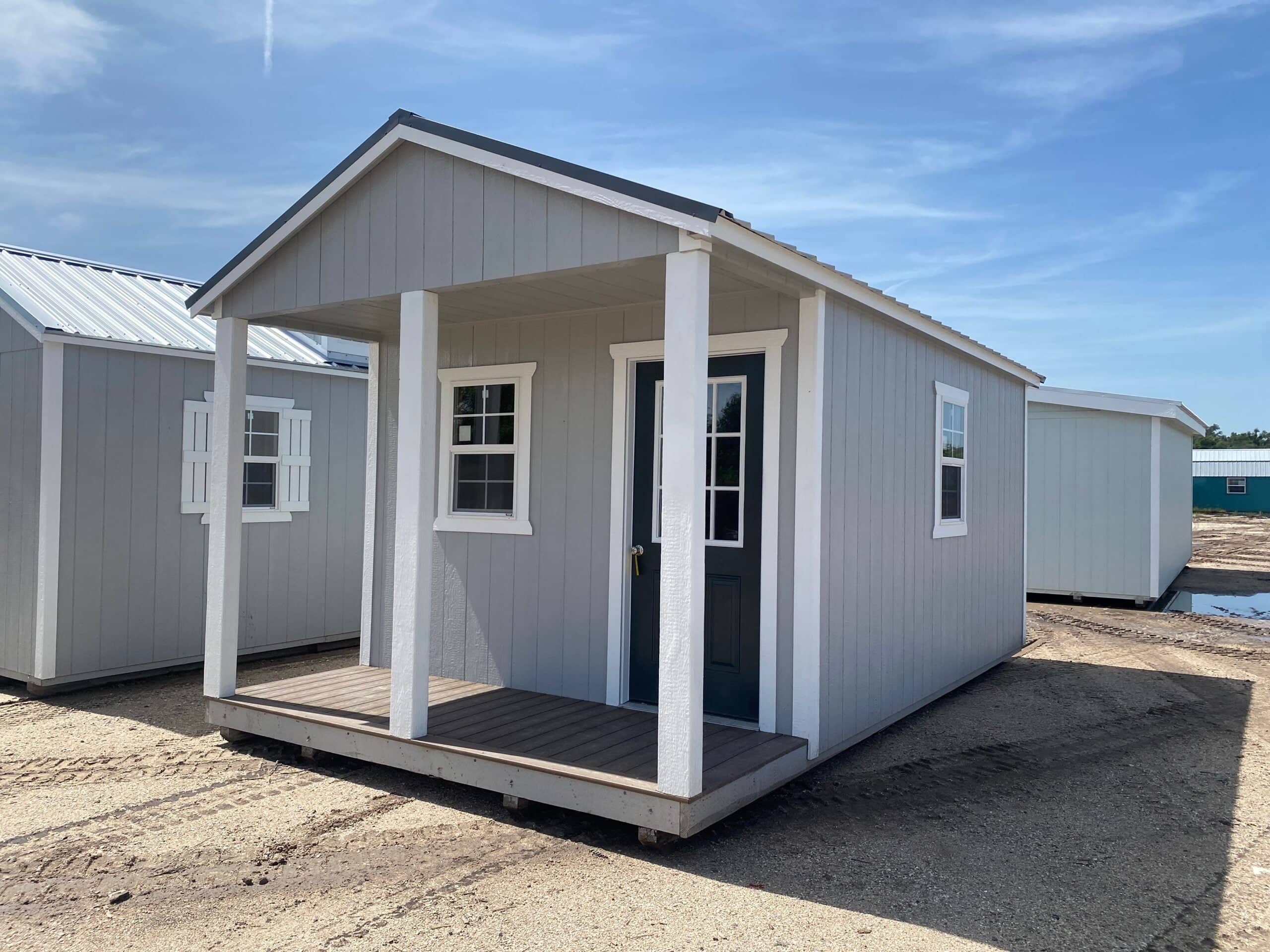 Light grey wooden cabin utility shed featuring white porch posts, dark green front door with glass grid windows, white trim, and a porch deck on a dirt gravel lot under a clear blue sky in Wildwood, FL.