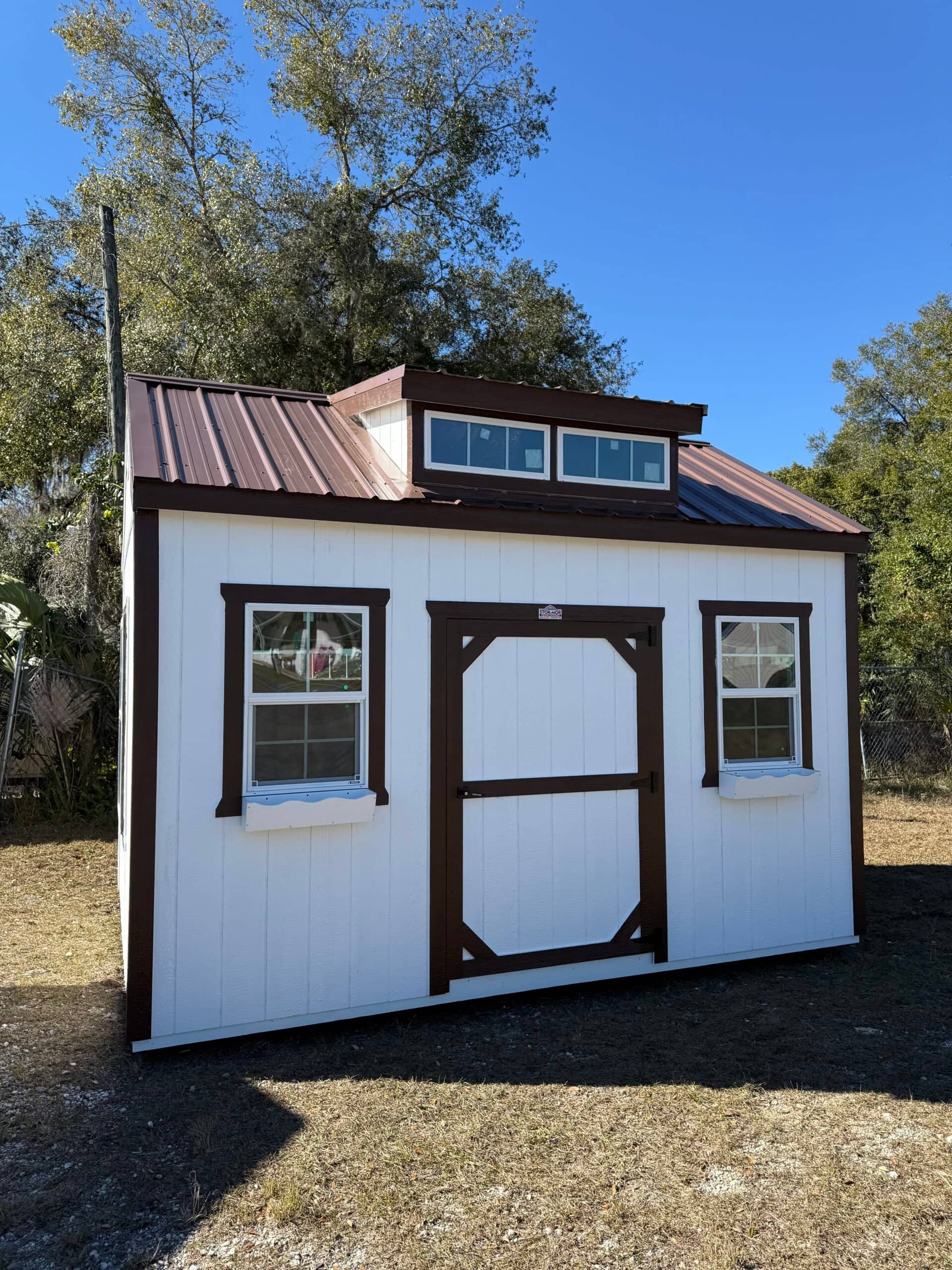 White wooden dormer studio shed featuring dark brown trim, a matching brown metal roof, top dormer transom windows, central front door, and flanking grid windows with flower boxes in Inverness, FL.