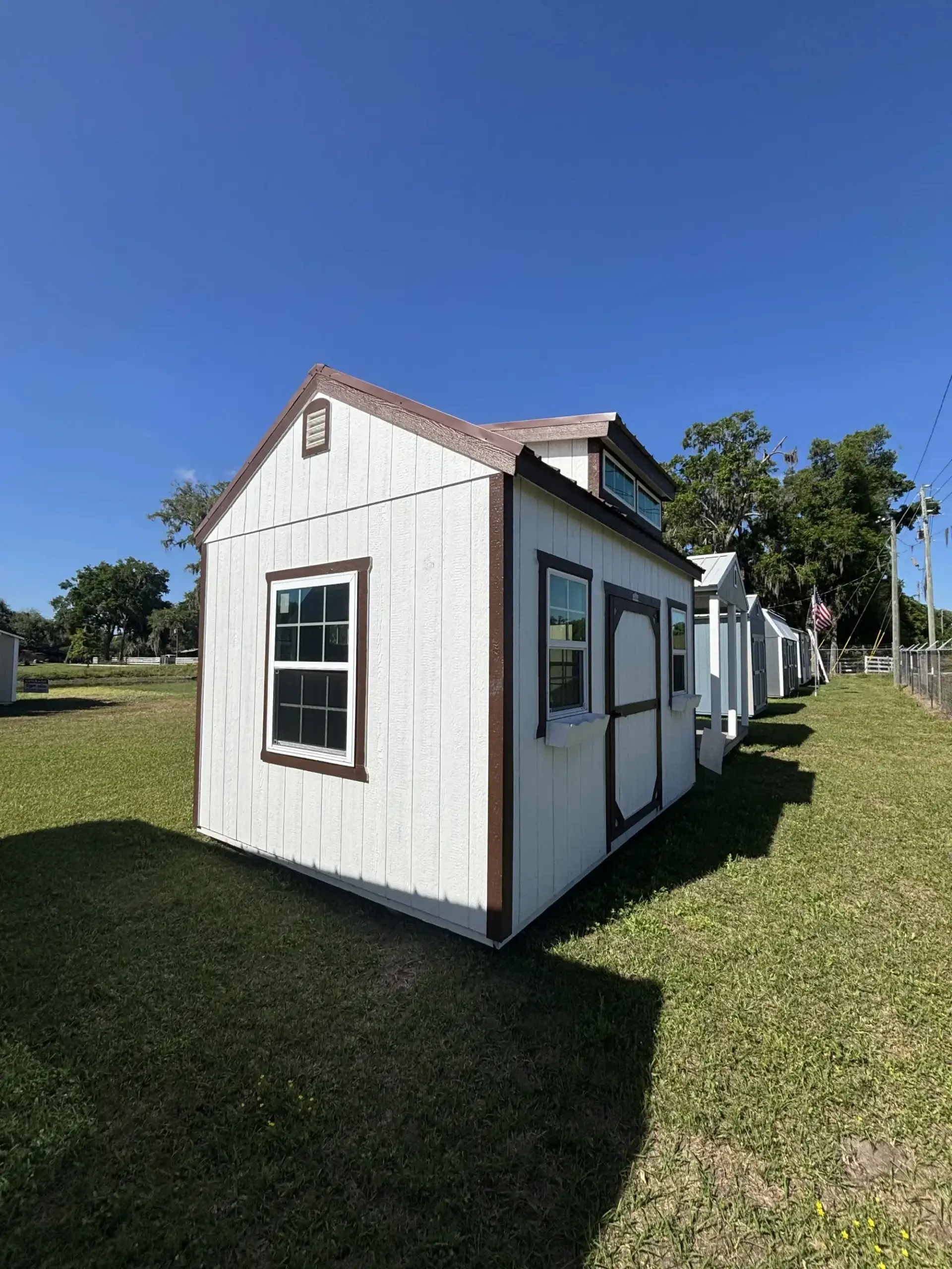 White wooden dormer studio shed with dark brown trim, grid windows, side entry door, and dormer windows sitting on grass in Leesburg, FL.