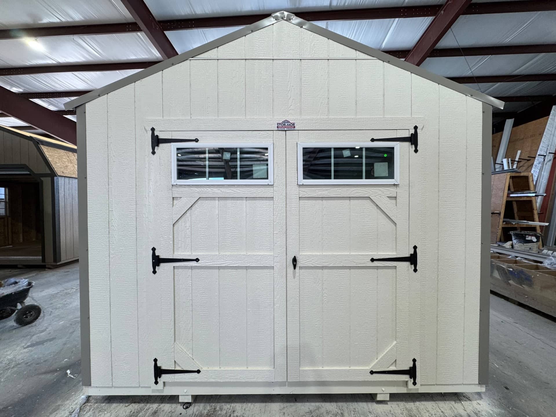 Off-white wooden gable utility shed featuring grey trim, double entry doors with top transom windows, and black T-hinges inside a workshop in Bushnell, FL.
