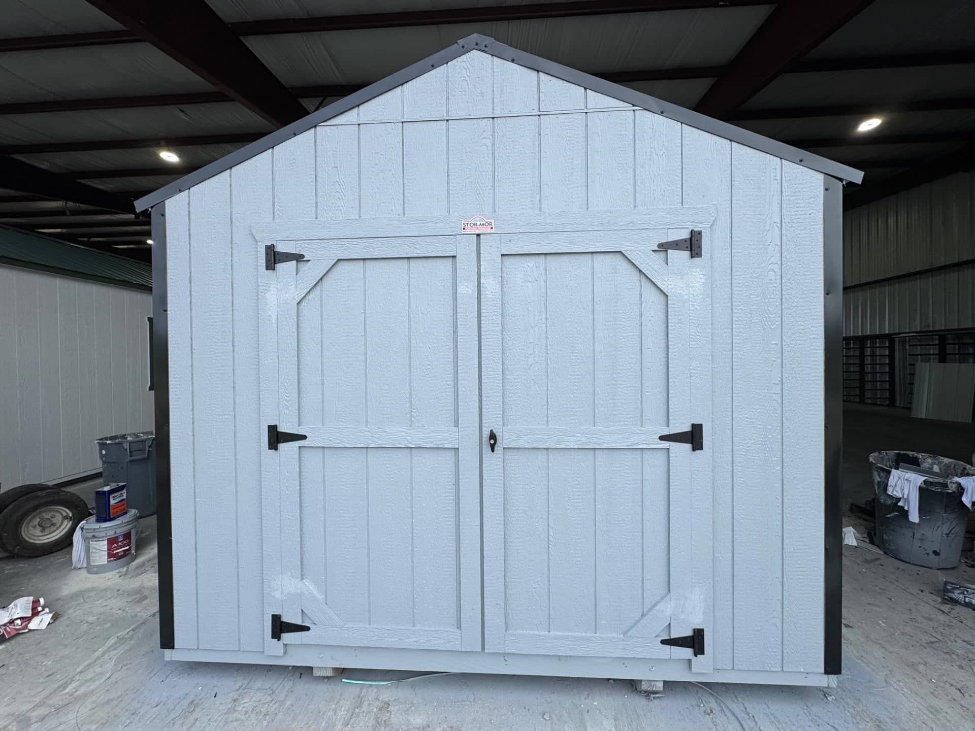 Light grey wooden gable utility shed with black corner trim, dark metal roof trim, double entry doors, and heavy-duty black hinges inside a workshop in Dunnellon, FL.