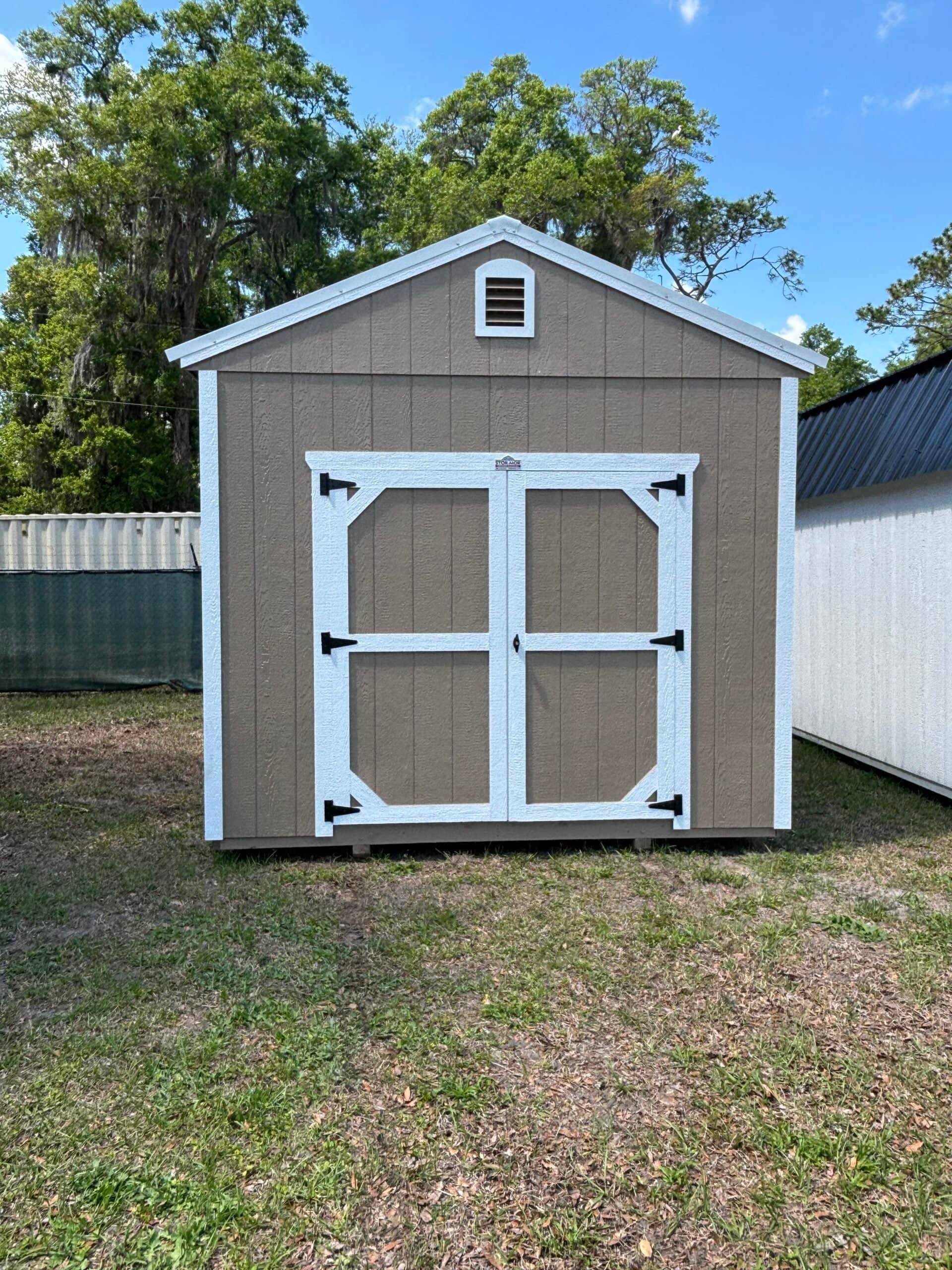 Tan wooden gable utility shed with white-bordered double front doors, white corner trim, and a top gable vent sitting on grass in High Springs, FL.