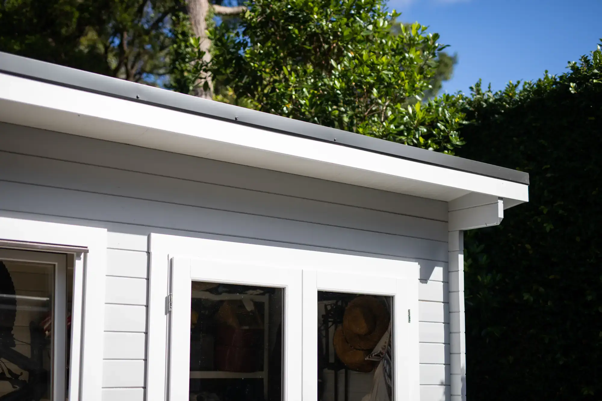 Close-up view of a white wooden garden studio shed with horizontal lap siding, white trim, glass panel french doors, dark roof edge, and lush green garden backdrop in Gainesville, FL.