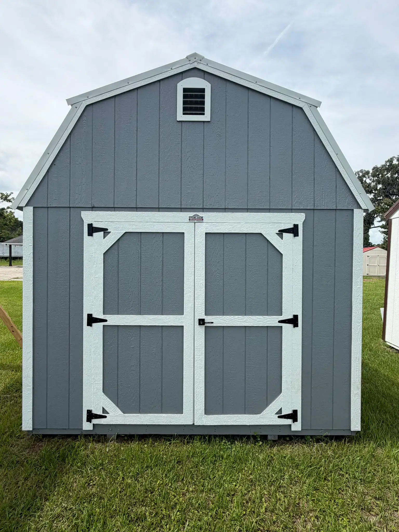 Dark gray wooden high barn shed with white trim, double front doors with black T-hinges, and an gable vent sitting on grass in Ocala, FL.
