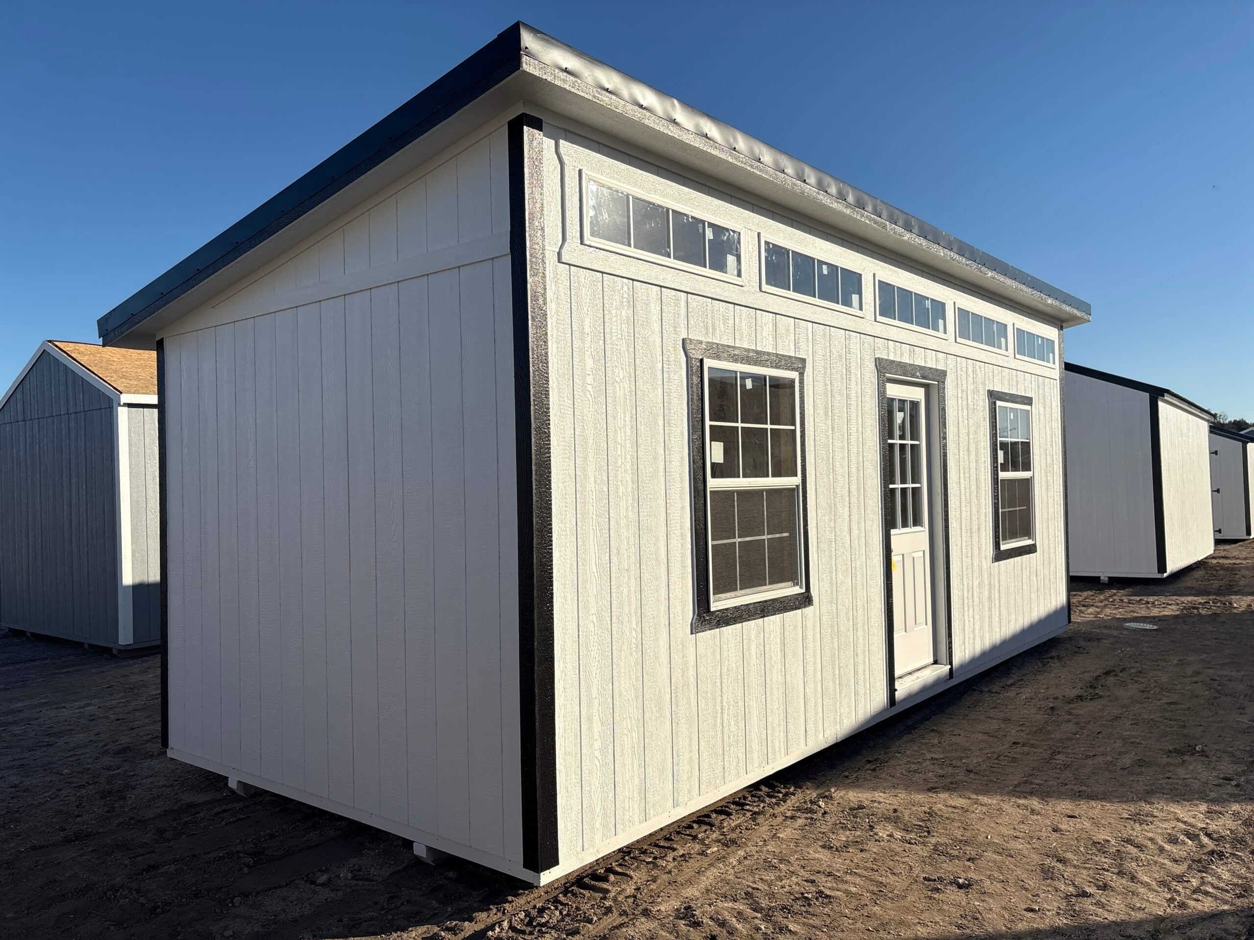 White wooden lean-to studio shed with black trim, upper clerestory transom windows, central door, and flanking grid windows sitting on a dirt lot in Leesburg, FL.