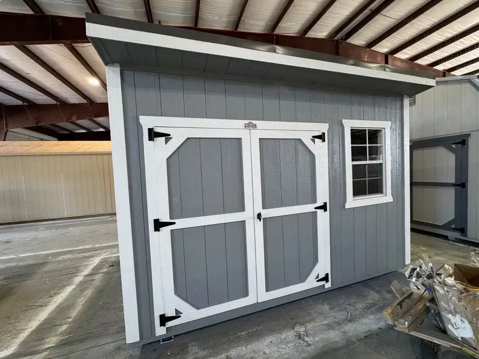 Grey wooden lean-to utility shed featuring white trim, white-bordered double center entry doors, a grid window, and a single-slope roof inside a workshop in Crystal River, FL.