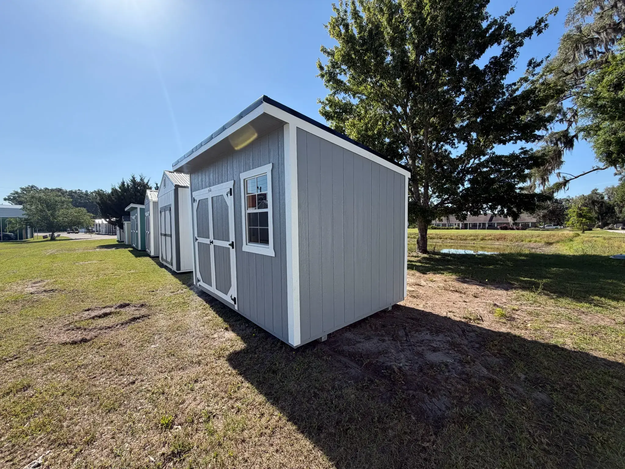 Grey wooden lean-to utility shed with white trim, white-bordered front double doors, a single grid window, and a single-slope roof sitting on grass in The Villages, FL.
