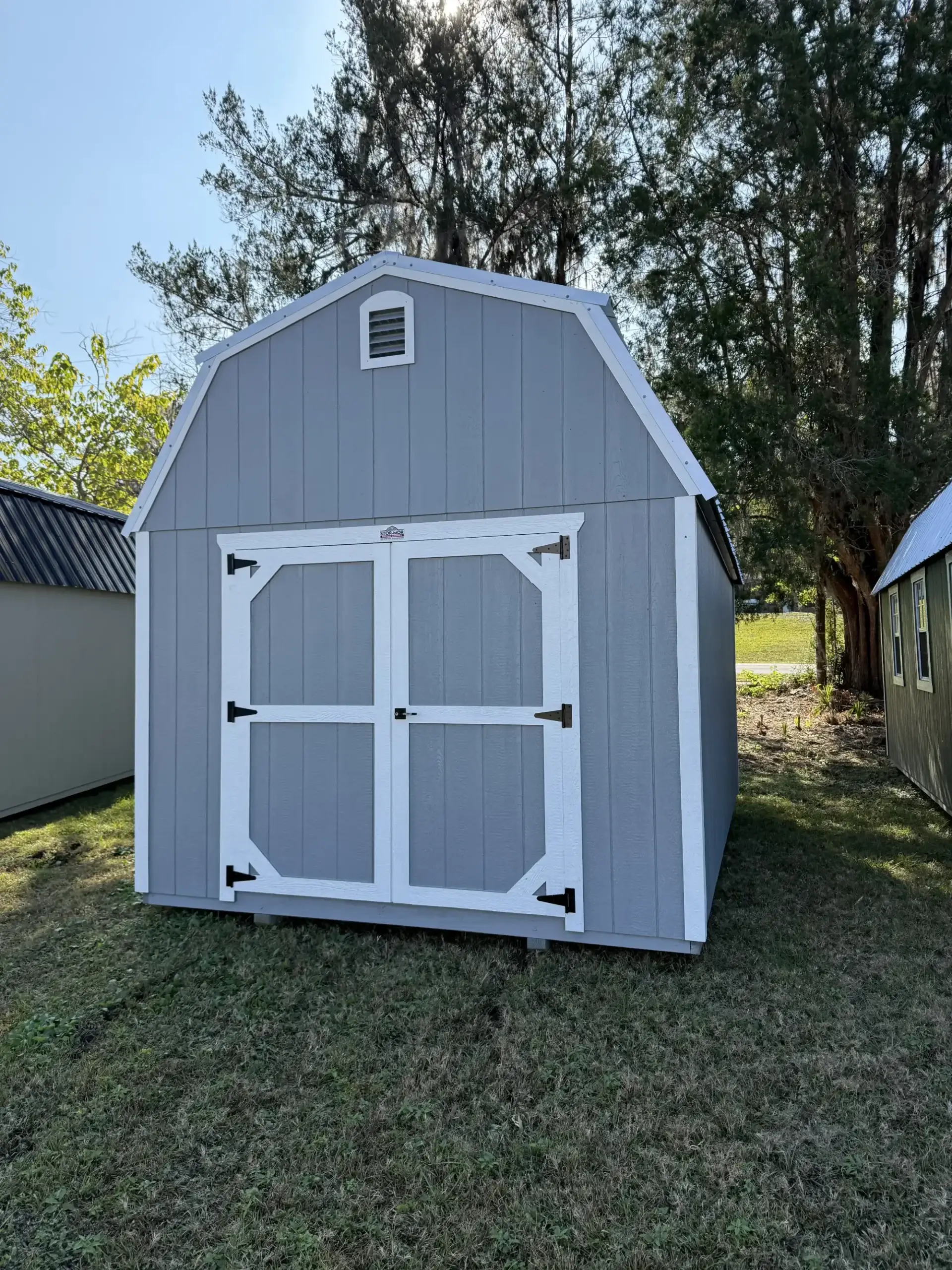 Grey wooden lofted barn storage shed with white trim, double entry doors with white cross bracing, and an upper gable vent in Brooksville, FL.