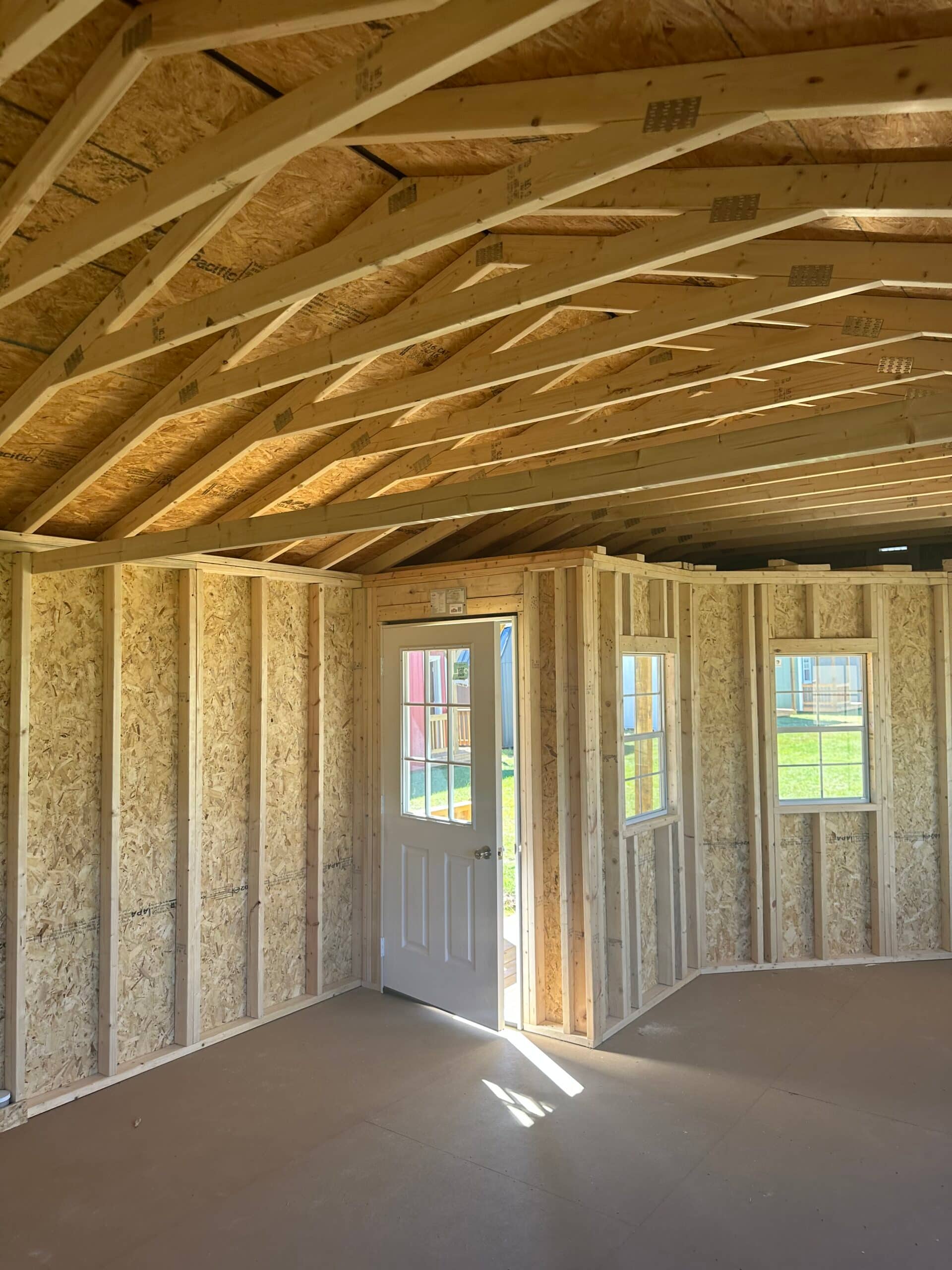 Interior view of unfinished custom wooden sheds framed with timber rafters, OSB wall sheathing, two windows, and an open glass-pane door in Ocala, FL.