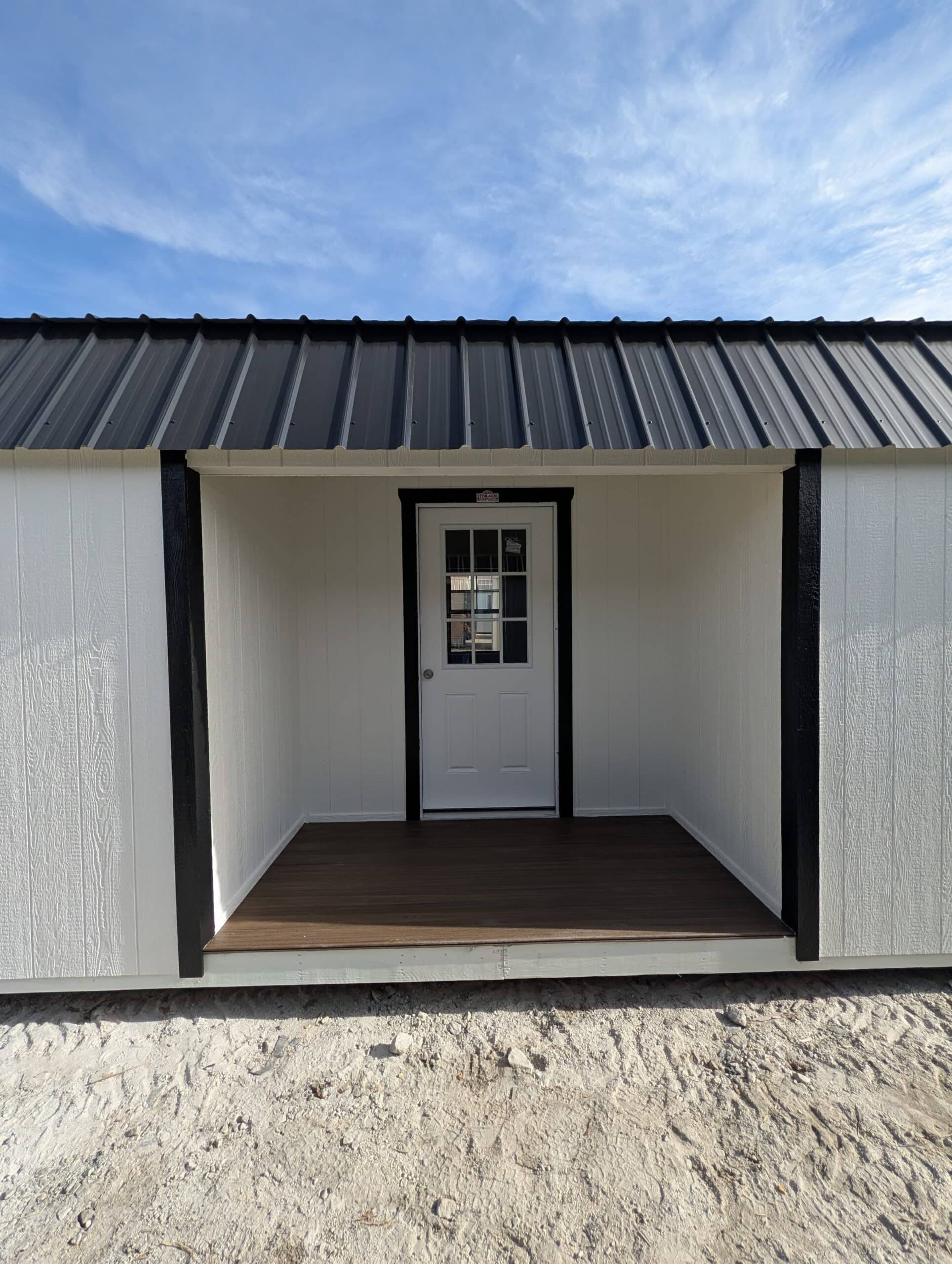 Close-up view of a white wooden side utility shed showing a covered porch entry, dark porch floor, bold black corner posts, white door with glass window grids, and black metal roof in Bronson, FL.