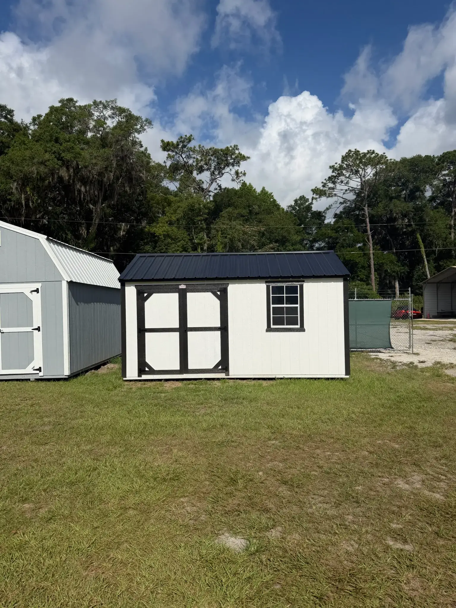 White wooden side utility shed with contrasting black trim, black double doors, a single grid window, and a dark metal gable roof sitting on grass in Bronson, FL.