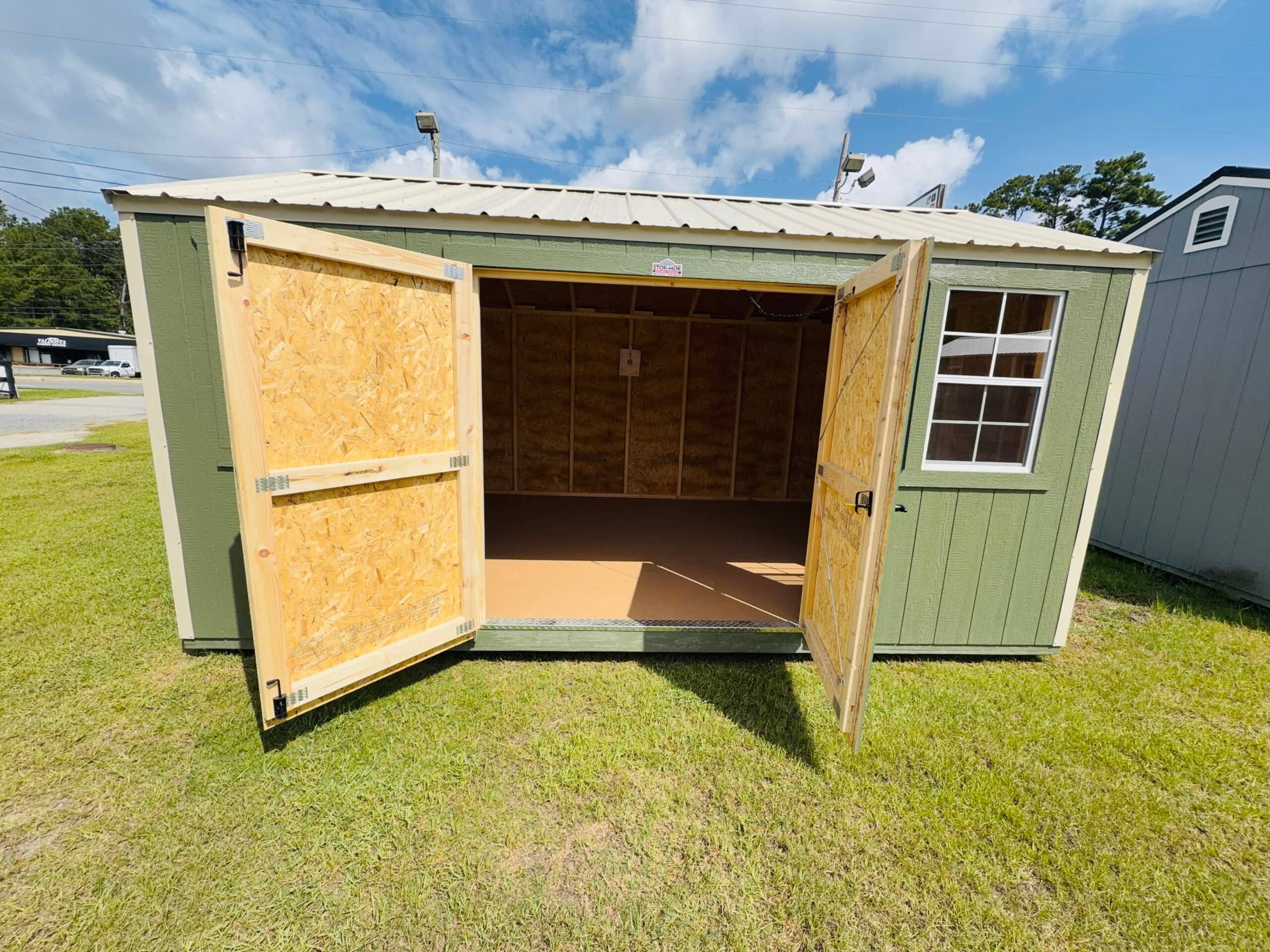 Sage green wooden side utility shed with tan trim, open double doors showing unfinished wooden interior, side grid window, and beige metal roof on grass in Chiefland, FL.