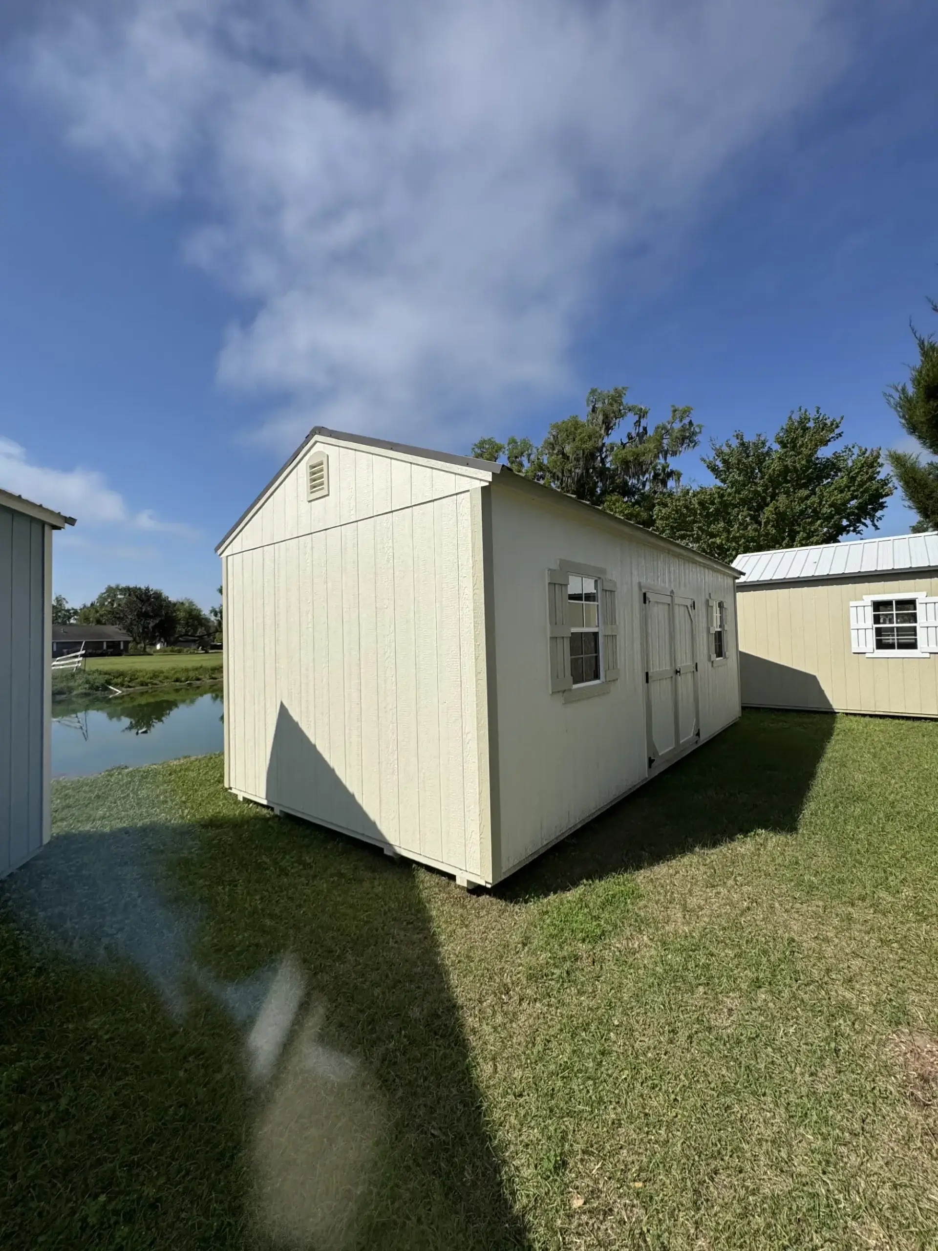 Long cream-colored wooden side utility shed with matching beige trim, central double doors, shutters around grid windows, and a gable vent on grass near water in Chiefland, FL.
