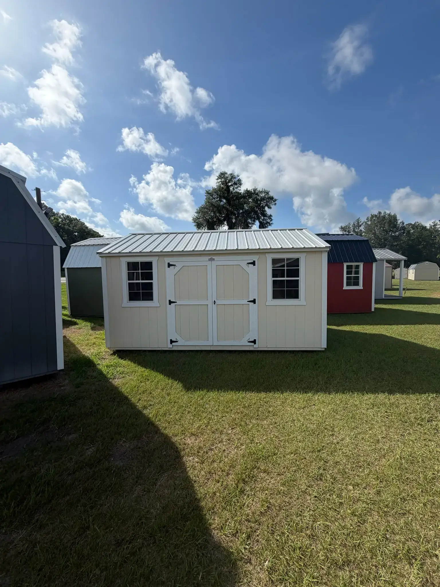 Cream-colored wooden side utility shed with crisp white trim, white-bordered double entry doors, two grid windows, and a silver metal gable roof sitting on a grass lot in Gainesville, FL.