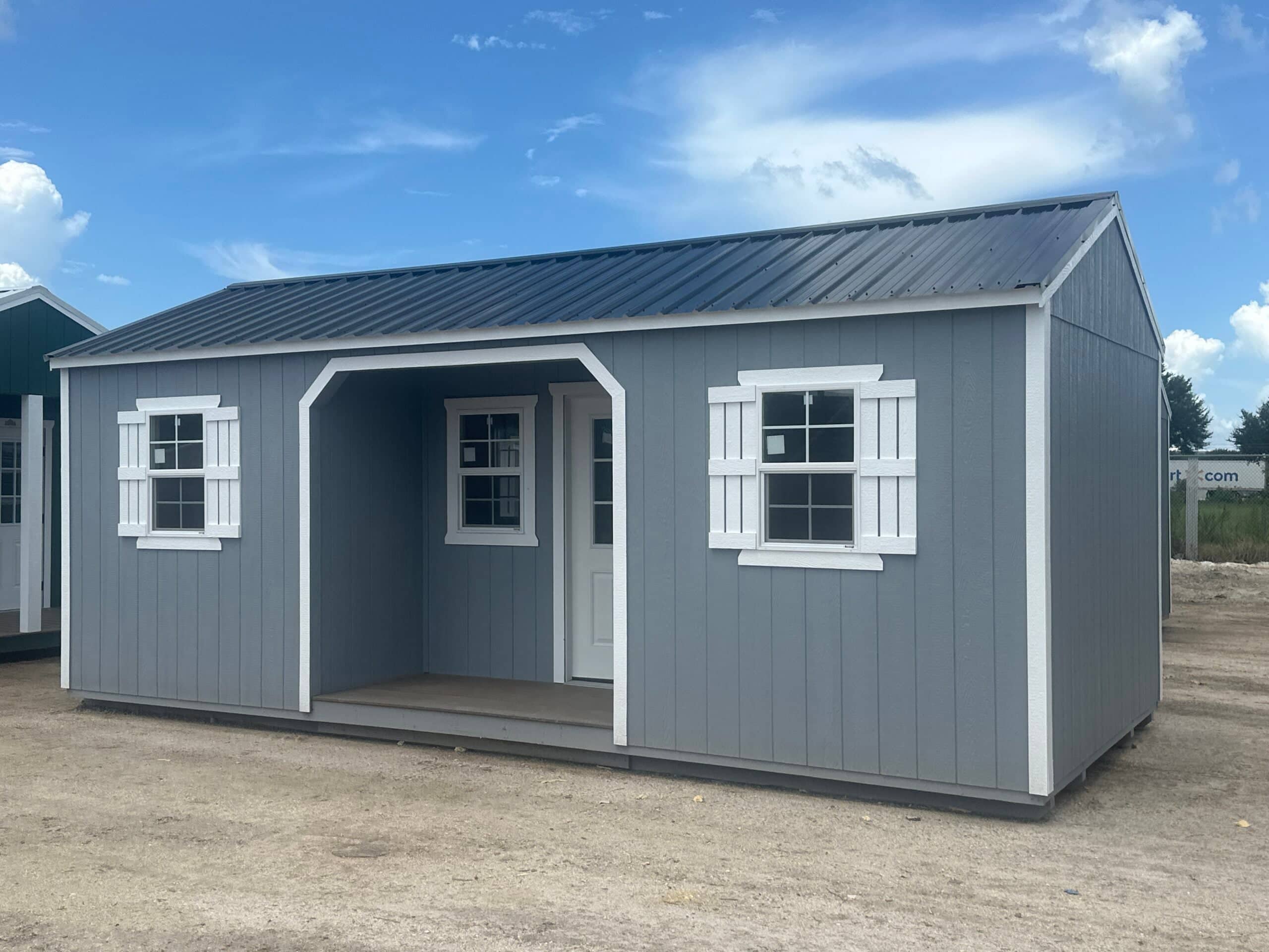 Slate blue wooden side utility shed with crisp white trim, decorative white window shutters, a white entry door, recessed porch alcove, and dark metal roof on dirt ground in Lecanto, FL.
