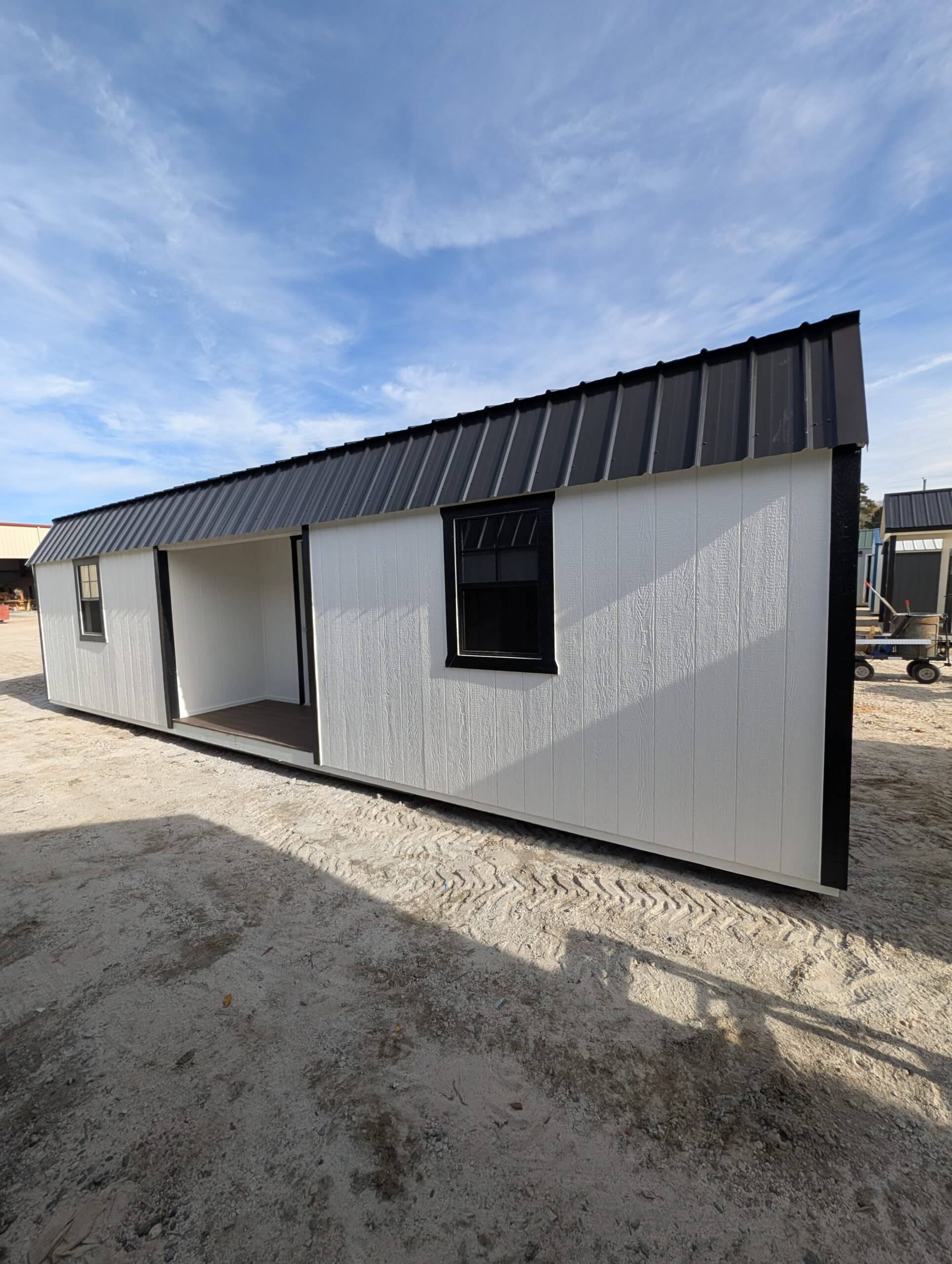 White wooden side utility shed featuring black trim, black framed windows, a recessed entry porch, and a dark black metal roof under a bright blue sky in Leesburg, FL.