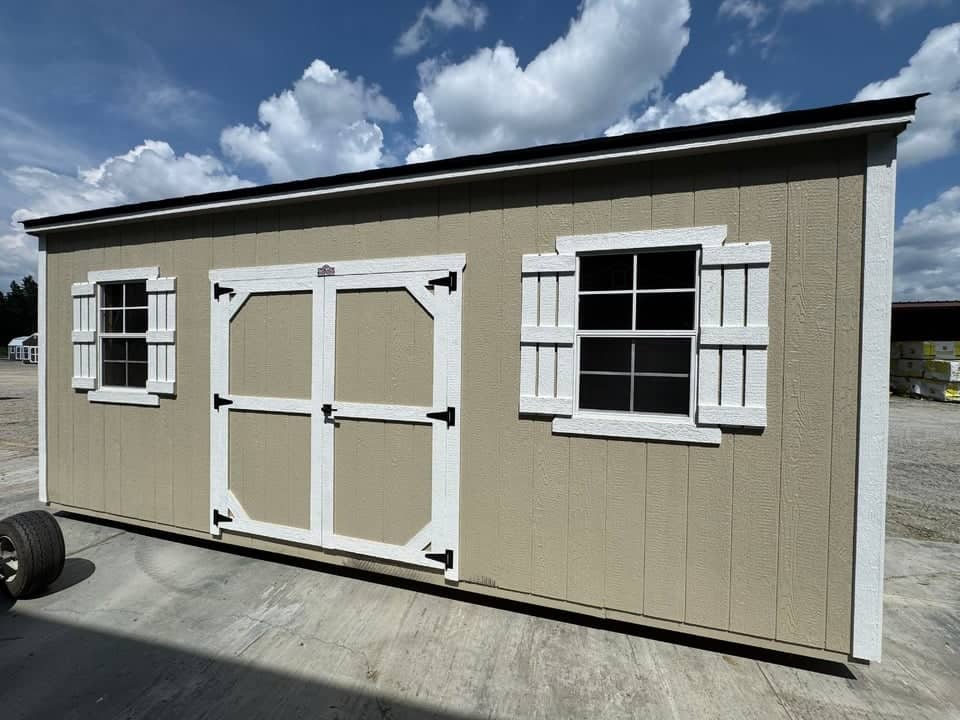 Tan wooden side utility storage shed featuring white trim, white window shutters, centered white double doors, and a dark roof line in Inverness, FL.