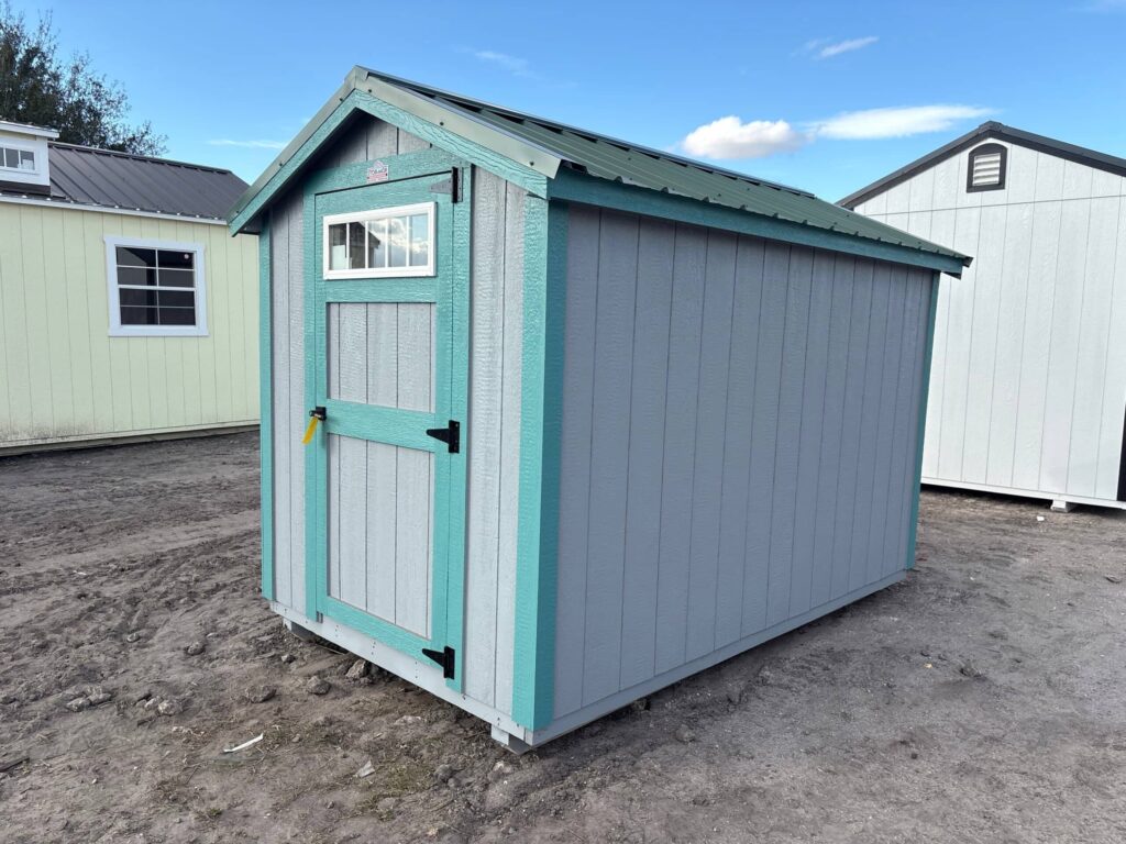 Compact grey wooden utility storage shed featuring teal accent trim, a green metal gable roof, and a single front door with an upper window pane sitting on a dirt lot.