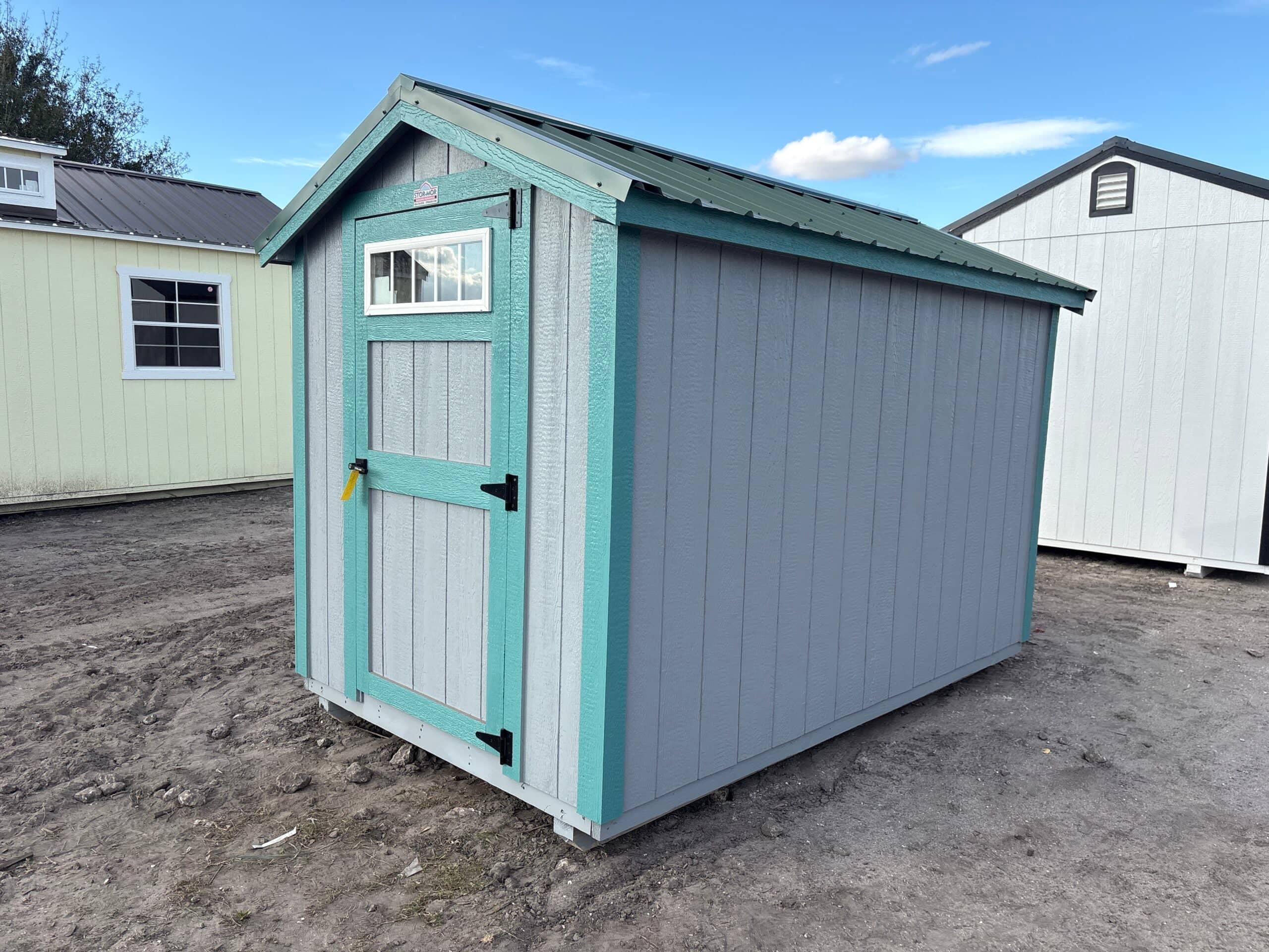 Compact grey wooden utility storage shed featuring teal accent trim, a green metal gable roof, and a single front door with an upper window pane sitting on a dirt lot in Spring Hill, FL.